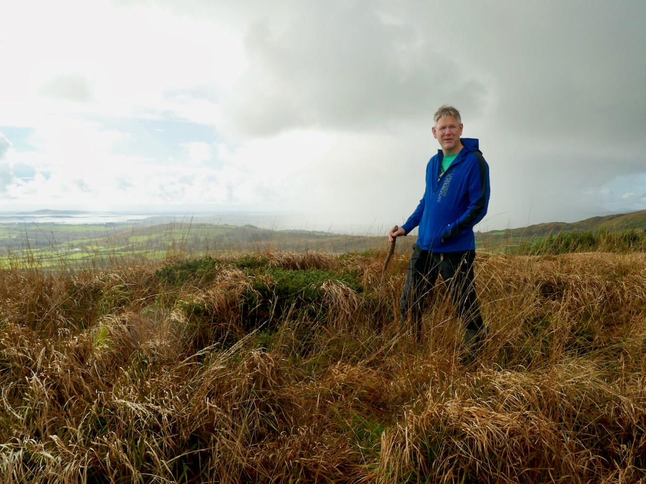Oliver standing on the mountain in a blue jacket with a walking stick, with rolling land and hills behind him